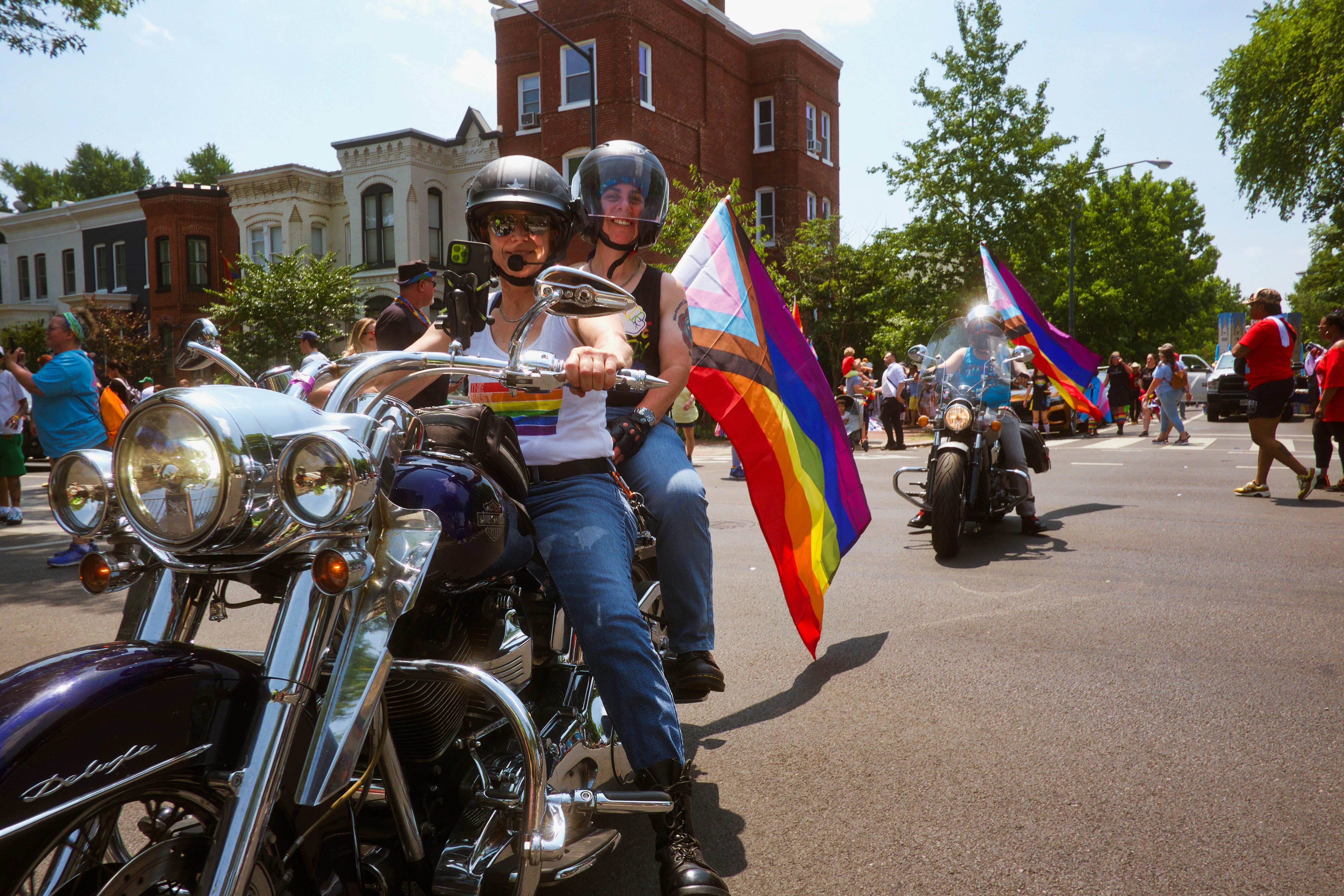 Two queer people smiling on the back of a motorcycle flying a trans-inclusive pride flag.