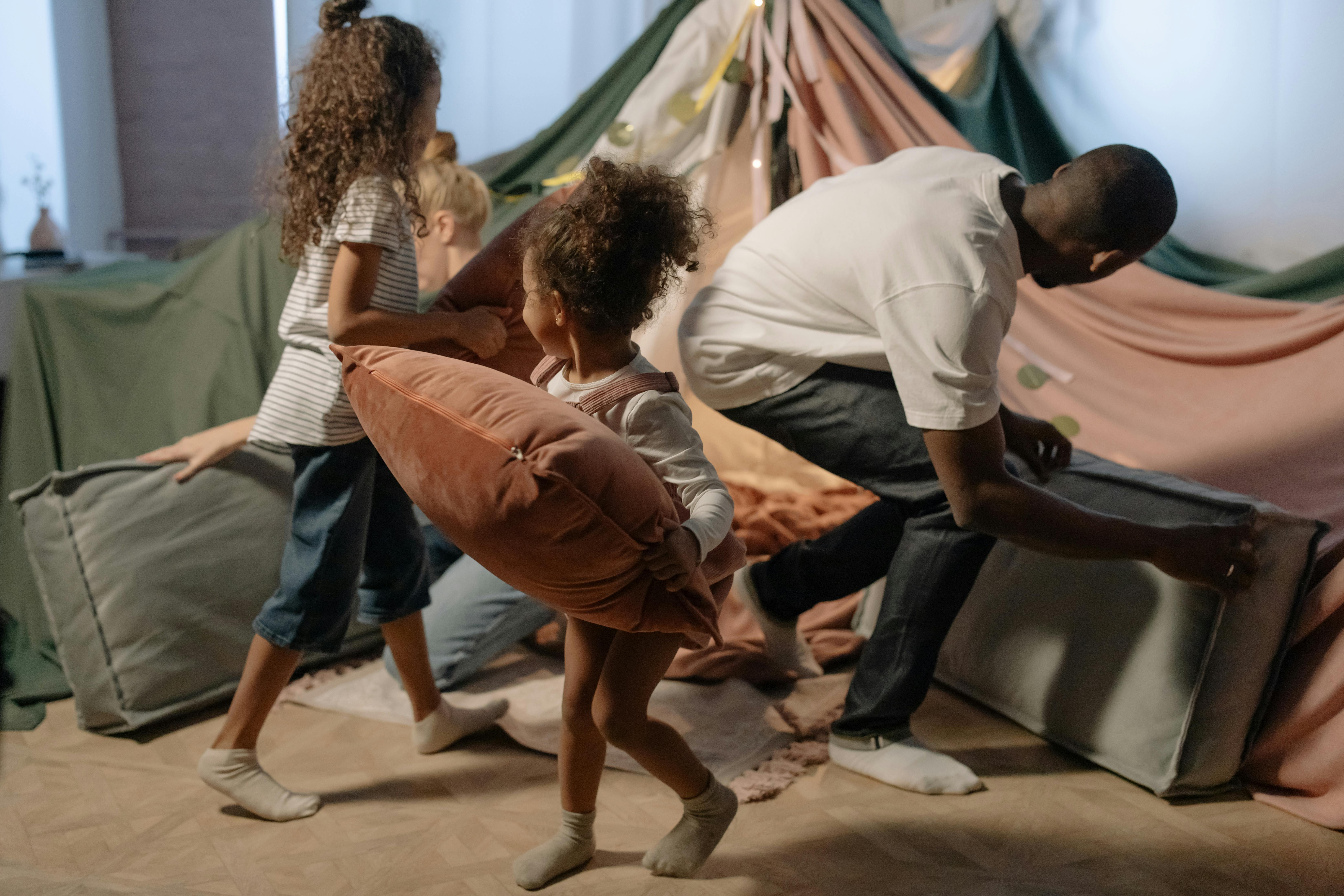 A young girl carries a cushion to build a pillow fort and blanket tent inside her cozy home with her sister, father, and mother.